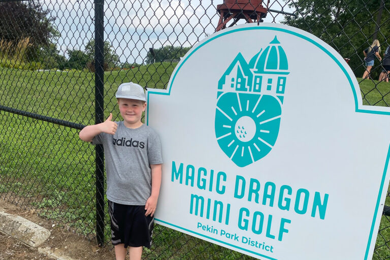 young boy giving a thumbs up in front of a Magic Dragon Mini Golf Sign